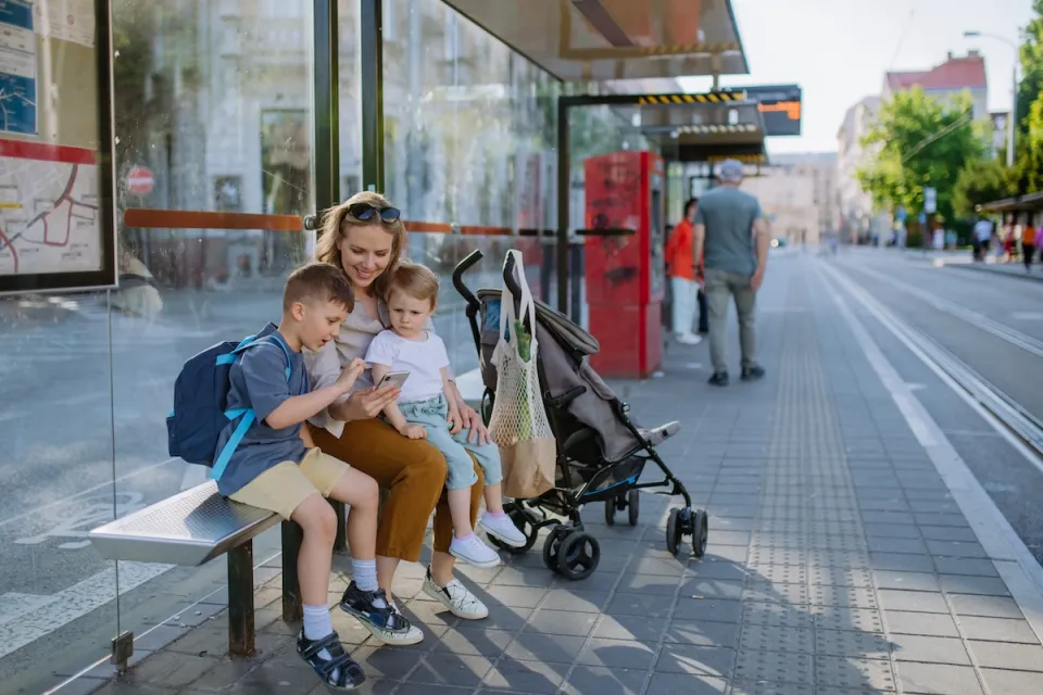 Woman and two children sit on bench at public transit stop waiting for tram with stroller next to them.